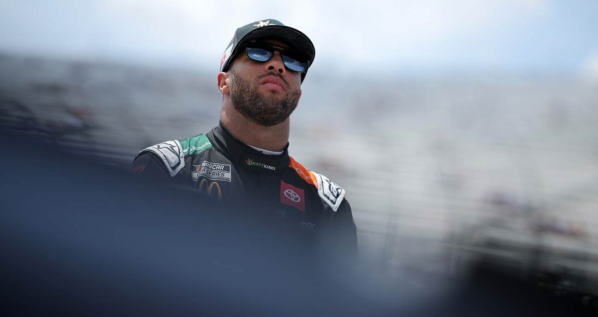 Bubba Wallace in sunglasses and a cap, looking up in front of the grandstands