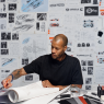 Heron Preston working at a desk in front of a wall of technical drawings