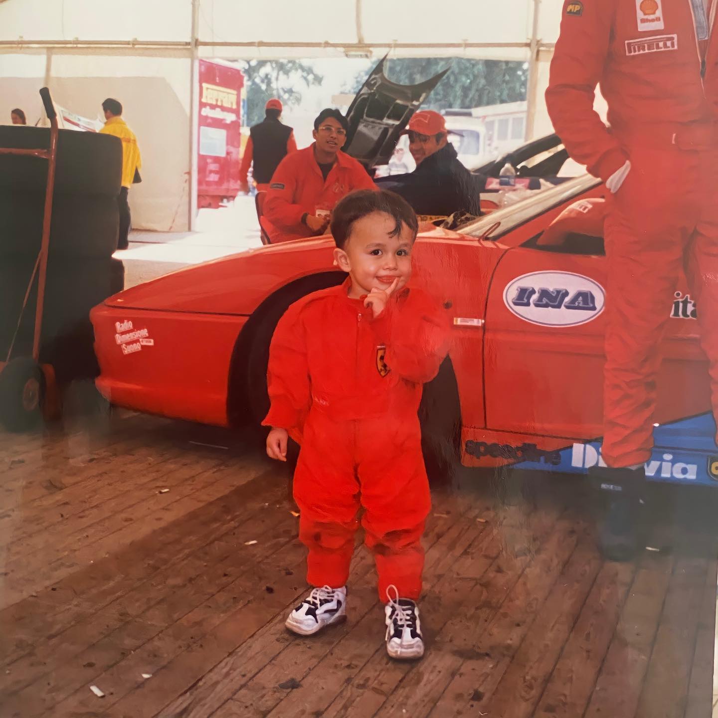 Alex Albon as a toddler in a red racing suit, standing in a paddock garage in front of a race car