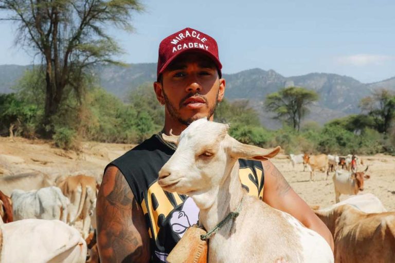 Lewis Hamilton in a red cap holding a white goat among a herd in a dry, hilly landscape