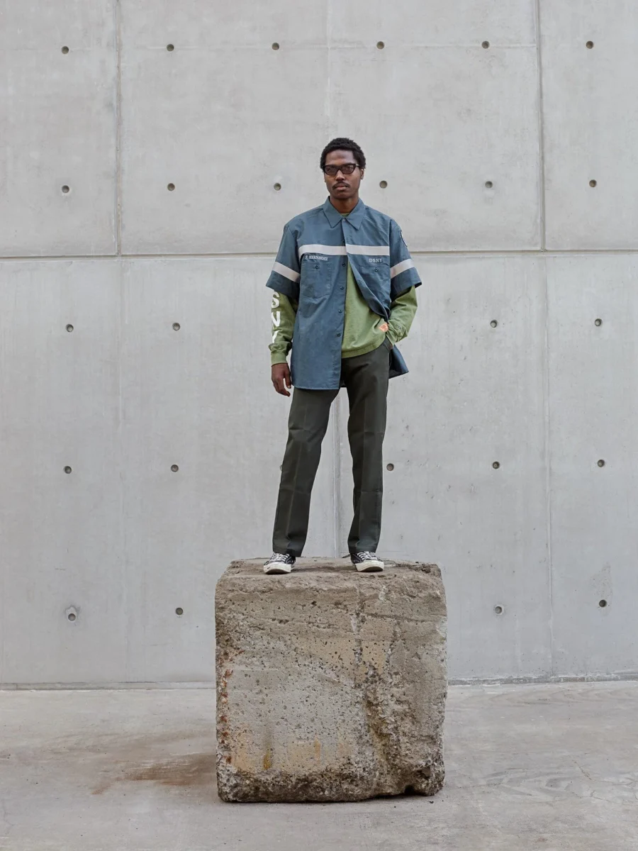 A model in a Heron Preston UNIFORM look, a reflective-striped DSNY shirt over a green layer, standing on a concrete block