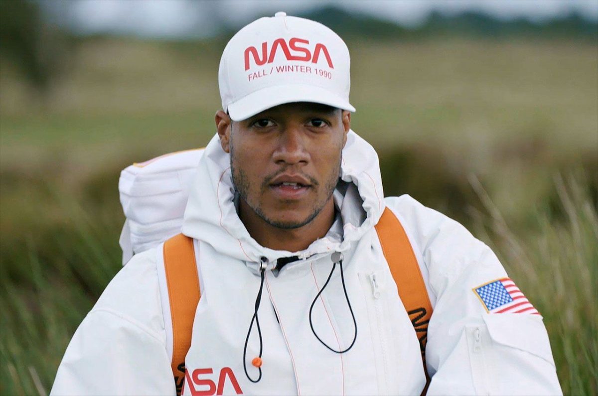 A man in a white NASA hoodie and cap with the worm logo, wearing an orange-strapped backpack